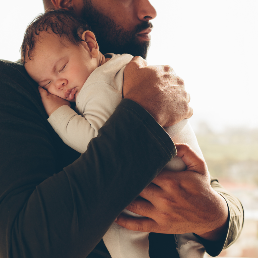 A Black man hugs a sleeping baby to his chest.